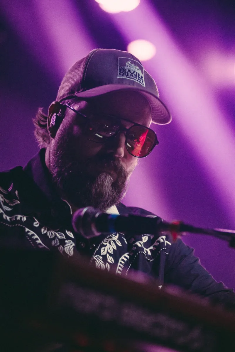 Big Holler member in a trucker hat and tinted aviators under purple stage light